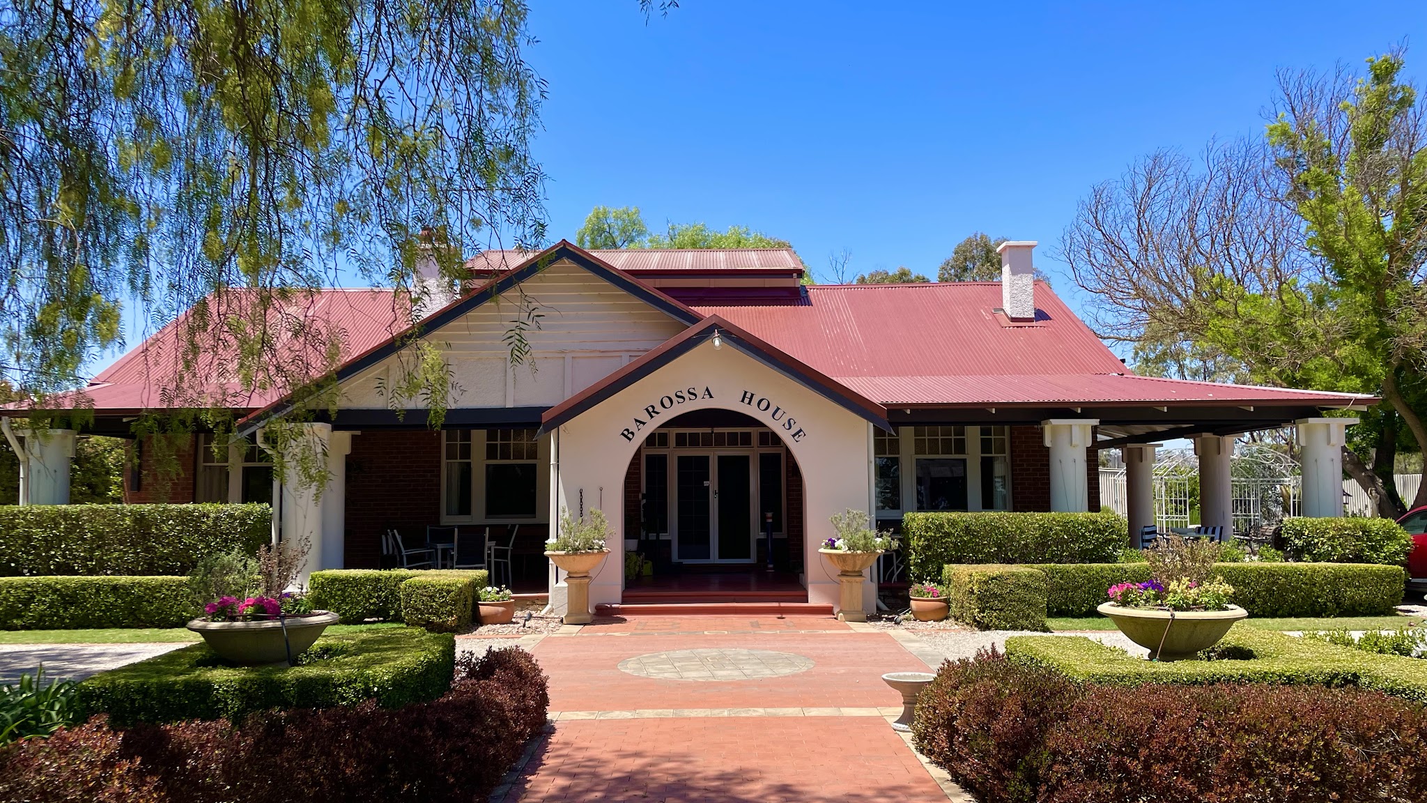 The front of Barossa House, a 1920s villa with a red roof and BAROSSA HOUSE painted on the entrance arch, behind a clipped box hedge garden