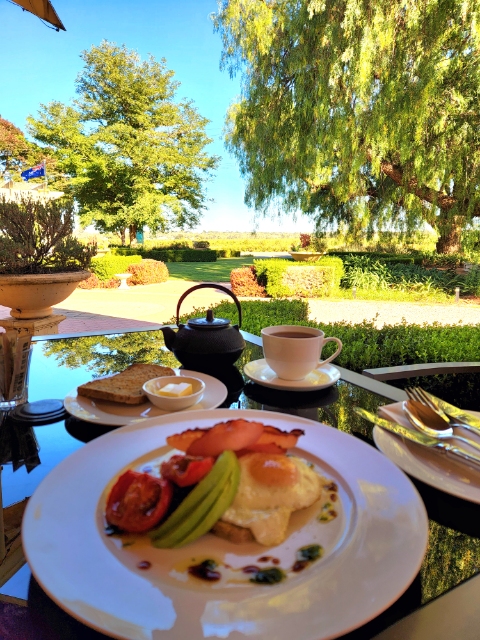 A cooked breakfast at Barossa House: egg on toast with bacon, grilled tomato and avocado, with coffee and a teapot, on a table looking out to the garden